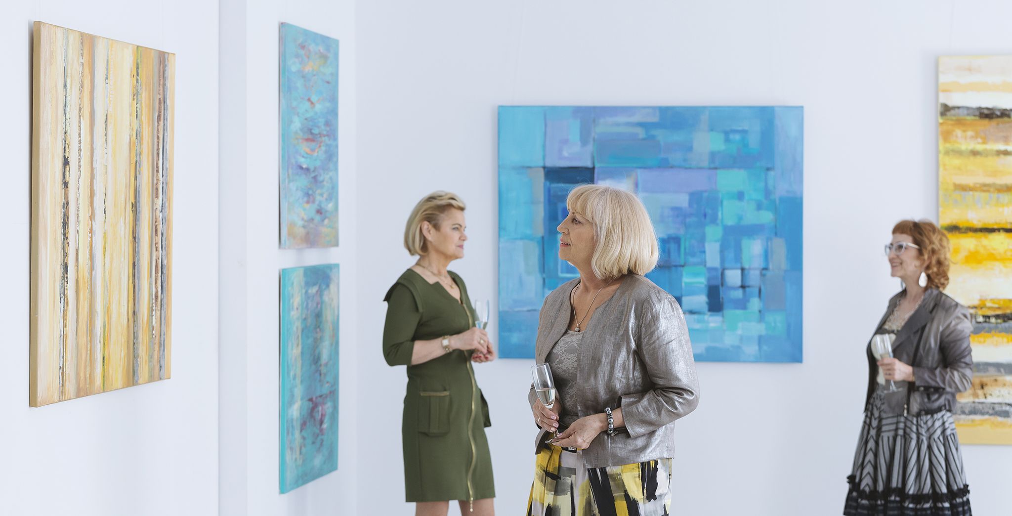 Three stylish older women enjoying an art gallery with drinks in hand