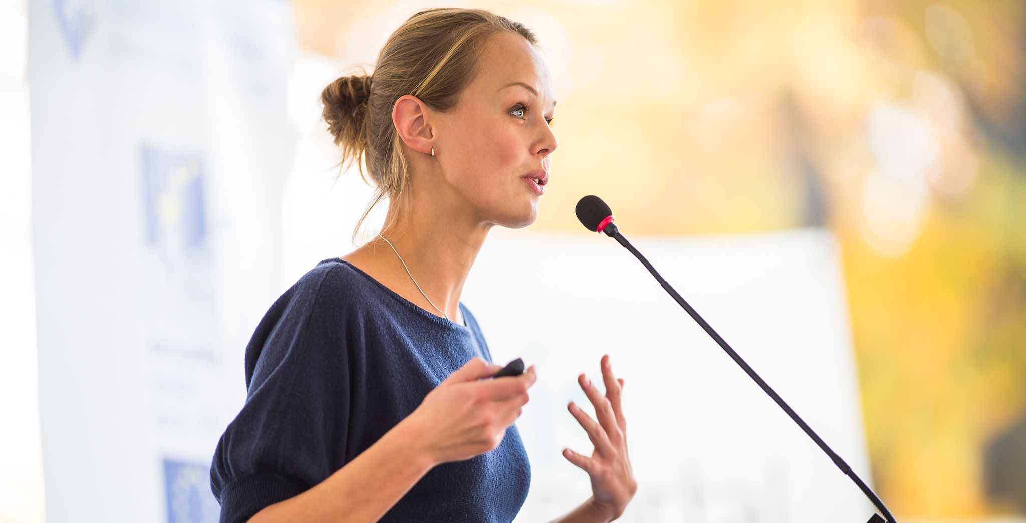 A blonde woman in a blue top speaks into a podium microphone